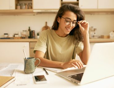 Charming Hispanic student girl in stylish glasses doing homework at kitchen table, learning distantly from home, using generic laptop. Attractive young female freelancer working on portable computer Charming Hispanic student girl in stylish glasses doing homework at kitchen table, learning distantly from home, using generic laptop. Attractive young female freelancer working on portable computer